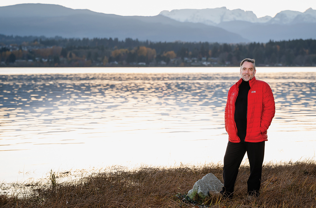 Dr Adam Thompson stands on a grassy shore in front of shimmering water, with trees and mountains in the distance.