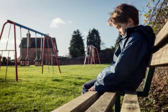 A child sits alone on a bench by a playground, looking down at the ground
