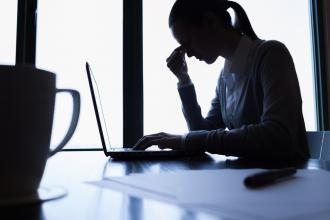 A woman sits in front of a laptop with her hand to her forehead