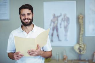 A chiropractor looks at a folder, with a spine model and diagrams behind him