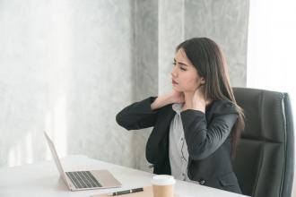 A woman sits at a computer with her hands on her neck and a look of pain on her face