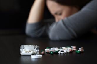 A young woman sits at a table, with her head down and an open bottle of pills in front of her