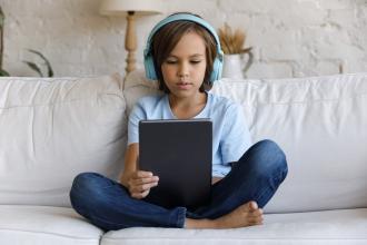 A child sits on a couch, looking at a tablet and wearing headphones