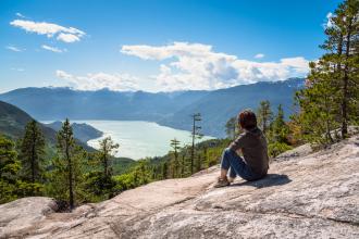 A person sits on the side of a mountain, looking at a lake below