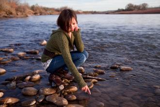 An Indigenous teen crouches down on a rocky shore, looking down, with her hand in the water