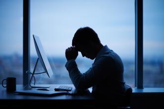 A person sits at a computer in an office, with their face down in their hands