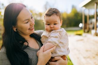 An Inuit woman holds a baby while standing outside in front of a building.