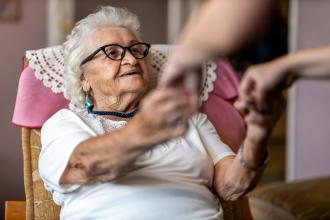 Female carer supporting old woman to stand up from an armchair at care home