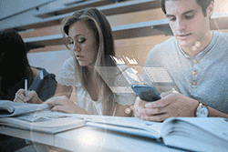 Students work at a desk; one is using a mobile phone