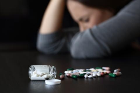 A young woman sits at a table, with her head down and an open bottle of pills in front of her