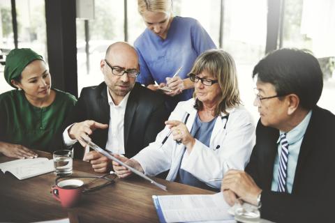 Five people are at a table, including a doctor and nurse, talking over a clipboard.