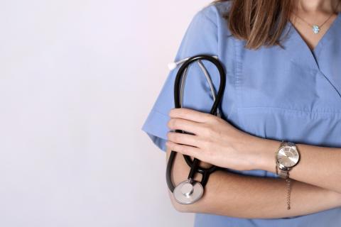 A doctor stands against a wall, holding a stethoscope