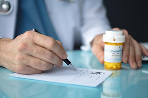 A doctor writes a prescription; a pill bottle is also seen beside the prescription paper