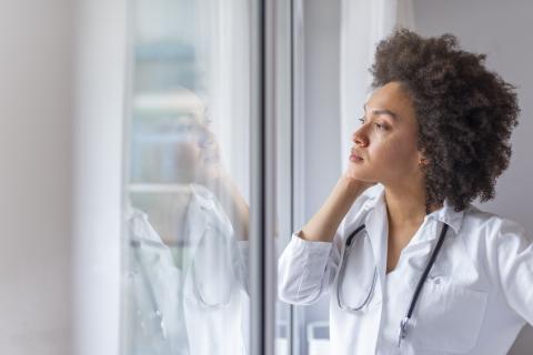 A young female doctor stands at a window, looking sad