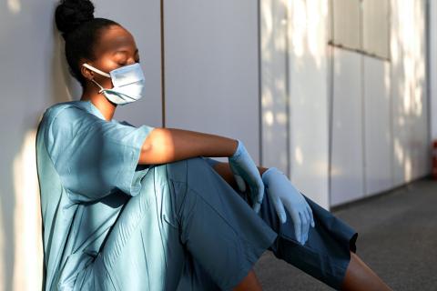 A doctor wearing a mask sits against a wall in a hallway, looking distressed and tired