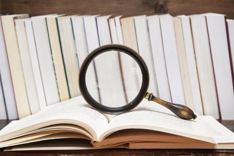 An open book with a magnifying glass on top, with a row of books on a shelf behind