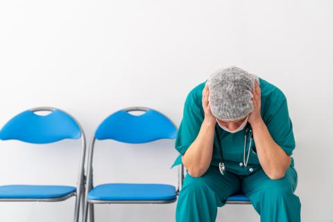 A doctor sits on a chair in a hospital waiting area with a mask on and his head in his hands