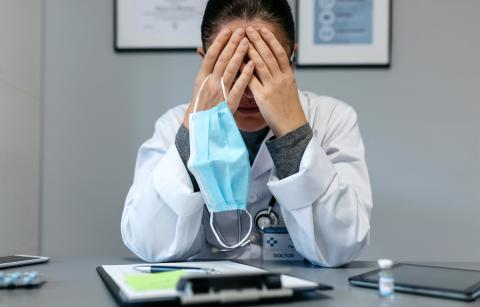 A doctor sits at a desk with their head in their hands, holding a face mask by the ear loop