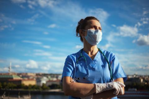 A doctor wearing a mask and gloves stands in front of a hospital, looking determined