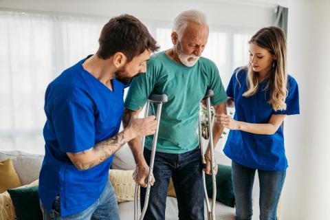 Two health care aides help a senior walk with crutches.