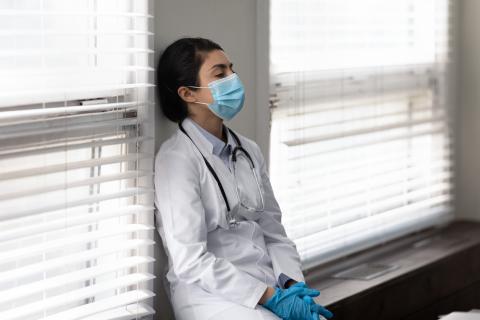 A doctor wearing a mask leans against a wall, looking exhausted