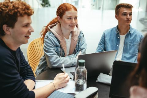 A group of teens sit at a table, talking; one has a laptop