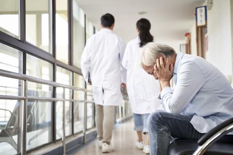 A man sits in a hospital waiting area with his head in his hands, as two doctors walk by in the background