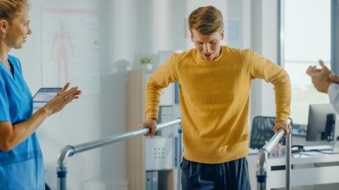 An injured worker in a physiotherapist's office walks while holding onto handrails.