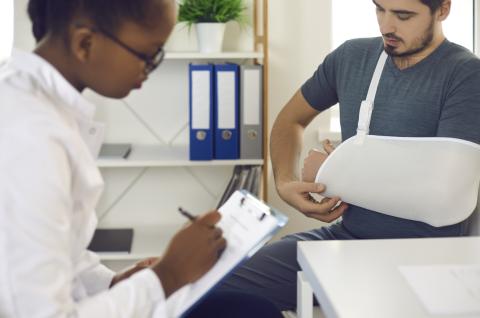 A doctor makes notes while consulting with a patient with their arm in a sling.