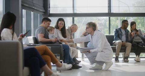 A doctor crouches down to talk to a baby sitting with its parents in a full waiting room.