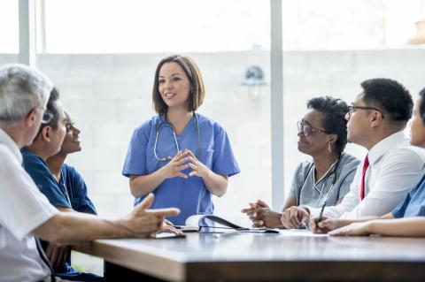 A doctor stands at one end of a boardroom table talking to other doctors who are sitting around the table.