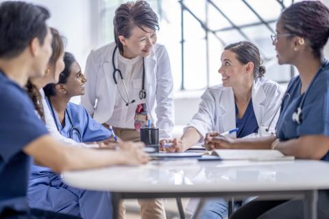 A smiling doctor stands at a table talking to a group of younger doctors.