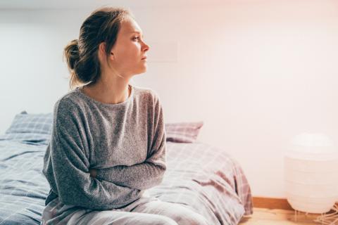 A woman sits on a bed, with her hands folded across her stomach.