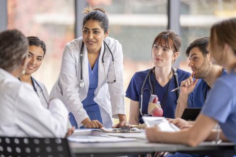 A group of doctors sit at a boardroom table, with one standing at the head of the table.