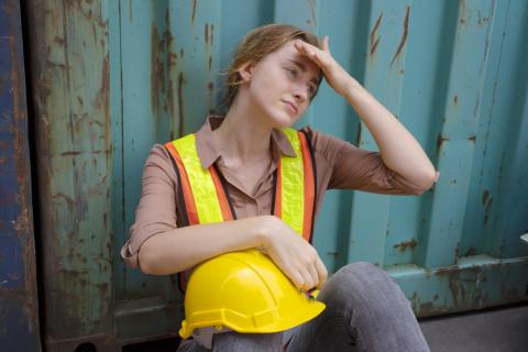 A person wearing high-visibility vest sits holding a hard hat on their lap, with their other hand to their forehead.