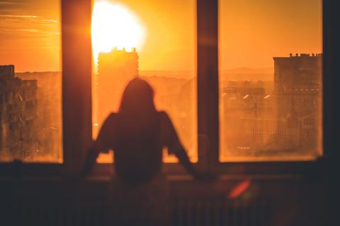  A woman stands at the window looking out at a bright red-orange sky with a glowing sun.