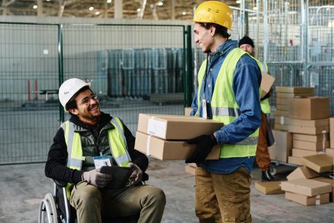 A warehouse worker in a wheelchair uses a tablet while talking to another warehouse worker holding a stack of boxes.