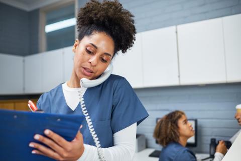 A medical office assistant talks on the phone while writing in a clipboard.