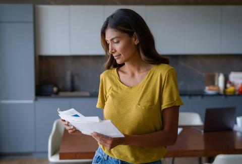 A woman stands in her kitchen looking through a stack of mail.