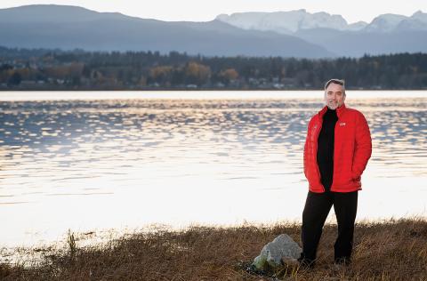 Dr Adam Thompson stands on a grassy shore in front of shimmering water, with trees and mountains in the distance.