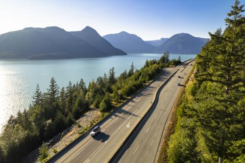 A coastal highway in British Columbia with a few cars, trees on either side, and water and mountains in the distance.