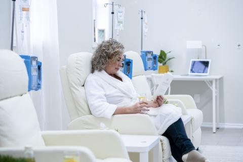 A woman wearing a bathrobe sits in a comfortable chair, reading and drinking a glass of water, while receiving IV therapy.