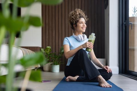 A woman sits on a yoga mat, drinking a smoothie and looking out the window.