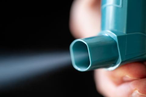 A hand is shown dispensing medication from an inhaler, with the mist visible in the air.