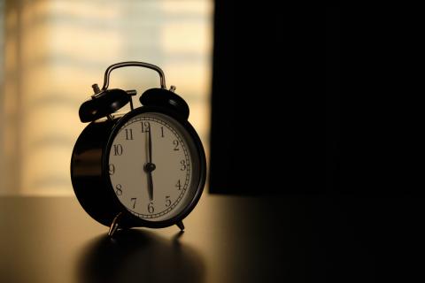 An alarm clock sits on a night stand in semi-darkness, indicating a time of 6:00.