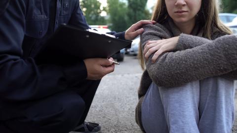 A police officer kneels down to offer comfort to a youth sitting on the ground with their arms around their knees.