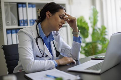 A doctor sits at her desk with her hand on her forehead, looking tired.