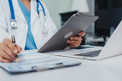 A doctor write on a clipboard while looking at a tablet and sitting in front of a laptop.