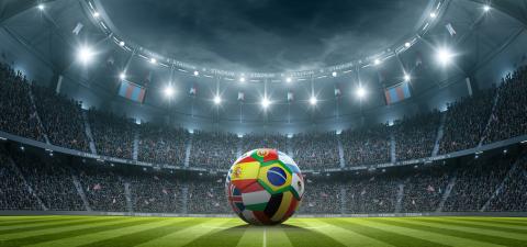 A soccer ball with international flags sits on a soccer field in an empty stadium.