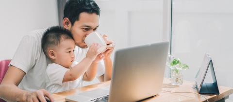 A dad and his young son sit at a computer while the son holds the dad's coffee cup up to the dad's mouth